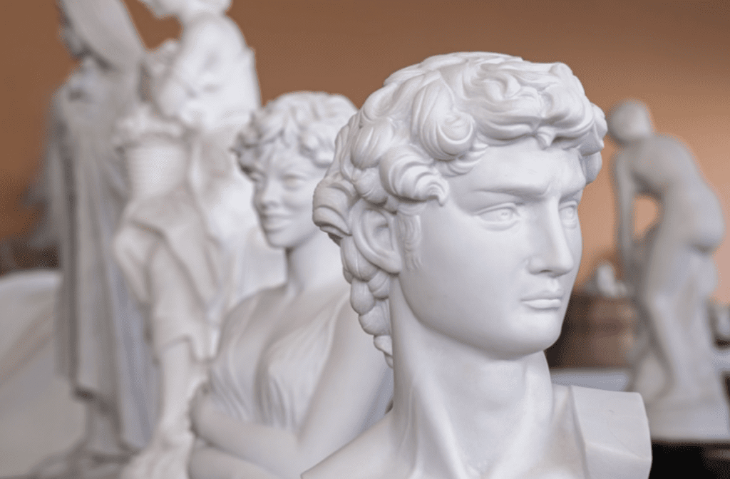 Close-up of a classical marble bust of a man with curly hair, showcasing timeless beauty. In the background, other sculptures, including a female figure, are blurred. The setting appears to be an art studio or gallery with a warm-toned wall.